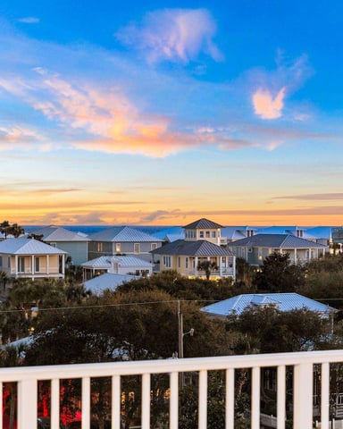 Coastal neighborhood at sunset with elevated homes, metal roofs, palm trees, and ocean view in distance