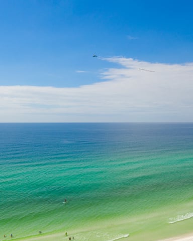 Sandy beach with turquoise water, white waves, swimmers, and colorful umbrellas under clear blue sky