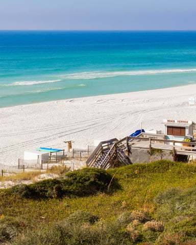 Sandy beach with turquoise water, wooden boardwalk, beach service station, and yellow flag, viewed from dune vegetation