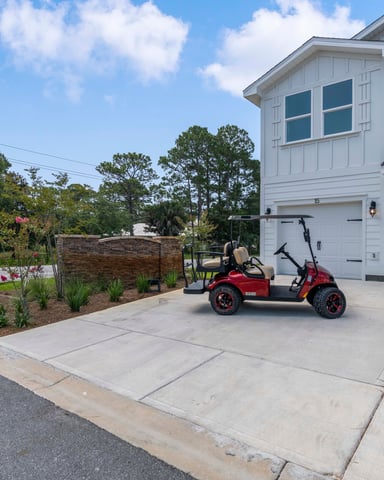 White two-story house with two-car garage and red golf cart parked on concrete driveway, landscaped front yard with trees and shrubs
