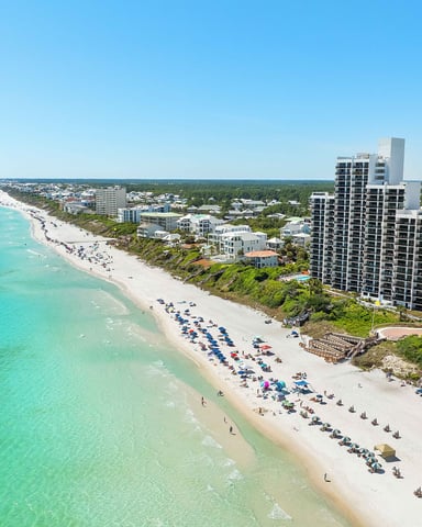 Aerial view of turquoise ocean, sandy beach with sunbathers and umbrellas, high-rise hotel, and residential buildings along tree-lined coastline
