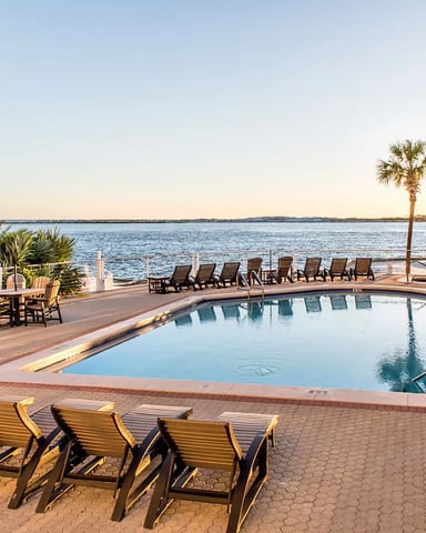 Beachfront resort pool with lounge chairs, palm trees, and bay view at sunset