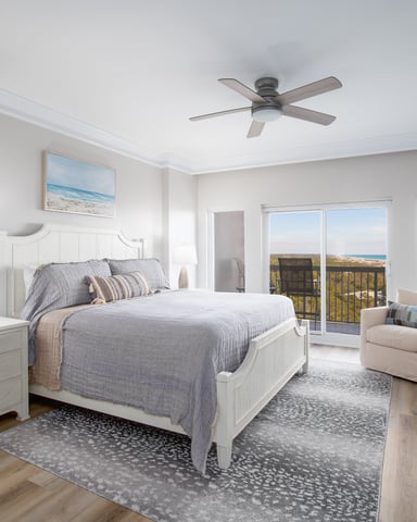 Bright coastal bedroom with white bed, ocean view from balcony doors, ceiling fan, and beach landscape artwork
