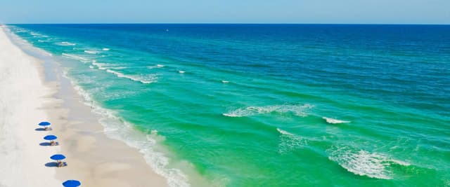 Aerial view of white sand beach with blue umbrellas and turquoise ocean waves under clear sky
