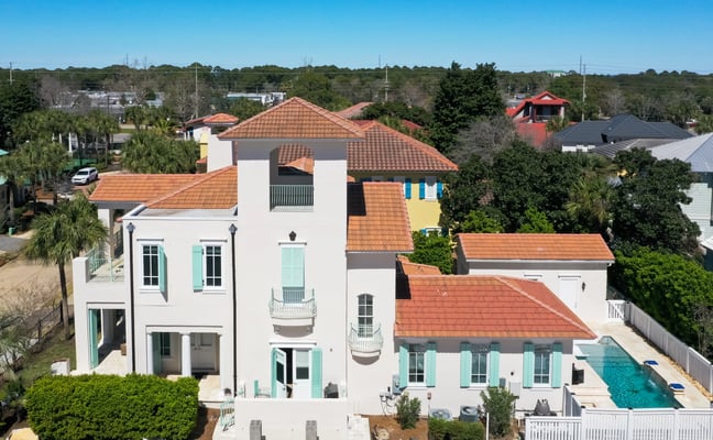 Aerial view of white Mediterranean-style home with terracotta tile roof, turquoise shutters, and backyard swimming pool surrounded by white fence