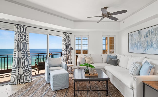 Bright beachfront living room with ocean view, white sectional sofa, patterned curtains, and balcony access