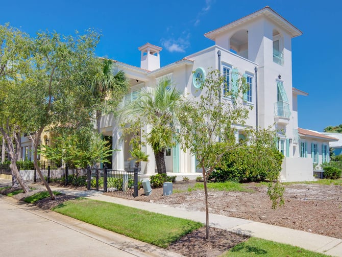 White two-story coastal house with tower, palm trees, and manicured landscaping under clear blue sky