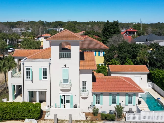 Aerial view of white Mediterranean-style home with terracotta tile roof, turquoise shutters, and backyard swimming pool surrounded by white fence